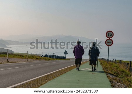 two people walking along the Cantabrian route near the beach of Meron, green pedestrian lane, traffic signs, meadows and cliffs in the background in a cloudless sunset.