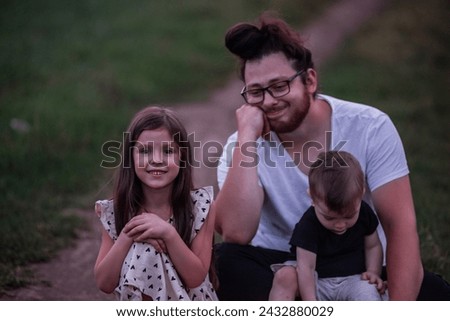 In the dimming light of dusk, father sits on rural trail, his children nestled in his arms, picture of protective love, quiet contemplation. Little girl and small boy playing near man