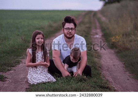 In the dimming light of dusk, father sits on rural trail, his children nestled in his arms, picture of protective love, quiet contemplation. Little girl and small boy playing near man
