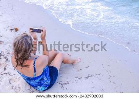 Woman in a bikini captures the beauty of the Atlantic Ocean landscape on her mobile phone while sitting on a sandy beach. Miami Beach. USA.