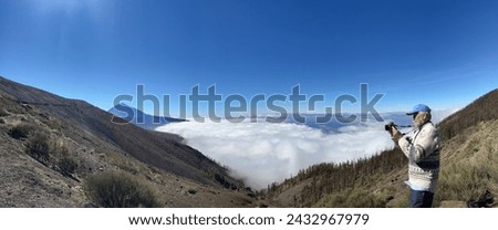 Panoramic view of bearded senior man tourist visiting the island of Tenerife takes photographs from the top of the mountain above a sea of clouds, in the distance rises the Teide volcano