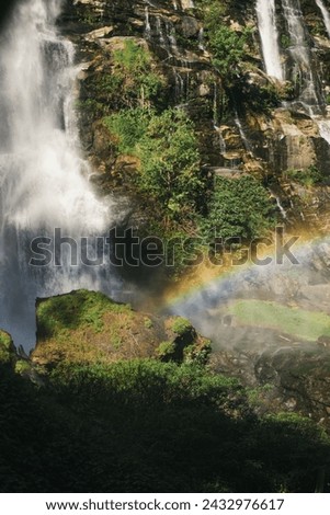 A cascading waterfall with a faint rainbow in the mist at the base. The surrounding area is lush and green.