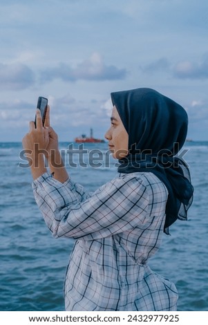 Asian Muslim woman taking photos using cellphone on the beach.