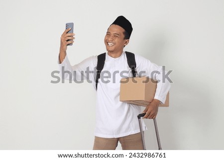 Portrait of excited Asian muslim man carrying cardboard box and suitcase while taking picture of himself or seflie and video calling. Going home for Eid Mubarak. Isolated image on white background