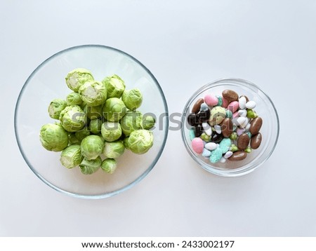 Fresh Brussels sprouts in glass bowl next to bowl of colorful candy on white background. Healthy versus indulgent food concept. Royalty-Free Stock Photo #2433002197