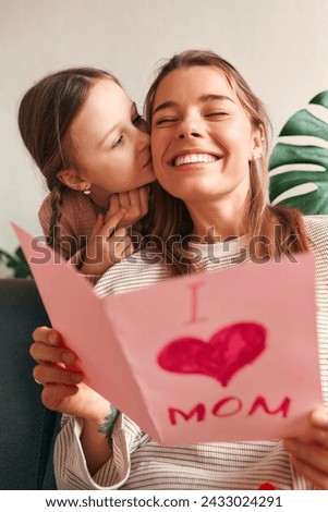 Child girl congratulating her mother by giving her a card while sitting on the sofa in the living room. Mother's day and women's day concept.