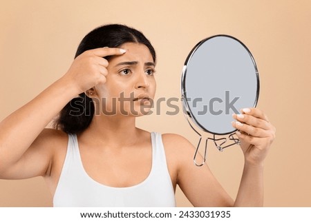 Concerned young indian woman closely inspecting her forehead in handheld mirror, worried female noticing pimple on face, suffering skin problems, standing against beige background, closeup Royalty-Free Stock Photo #2433031935