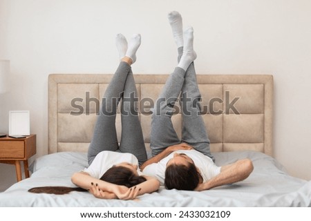 Couple enjoys playful moment in cozy bedroom, lying on their backs on the bed with legs up in the air, dressed in casual home wear, creating relaxed and cheerful atmosphere Royalty-Free Stock Photo #2433032109