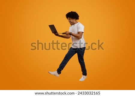 Excited curly millennial african american guy jumping up in the air with computer laptop in his hands, got job offer, reading great news, orange studio background, full length, copy space Royalty-Free Stock Photo #2433032165
