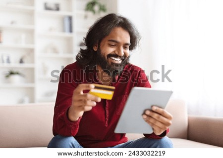 Cheerful smiling handsome young indian man sitting on couch in living room, using digital tablet and bank credit card, order food online, shopping on Internet from home, copy space Royalty-Free Stock Photo #2433032219