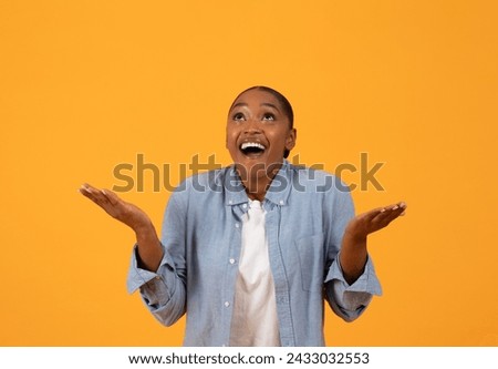Excited African American woman in blue shirt celebrates special offer with emotional expression, looking up and holding open palms, posing over orange studio backdrop. Advertisement banner