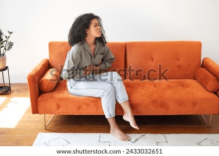 Gastrointestinal Distress. Young brazilian woman at home dealing with abdominal discomfort, touching aching belly, showcasing challenges of digestive issues and period pain, sitting on couch indoor Royalty-Free Stock Photo #2433032651