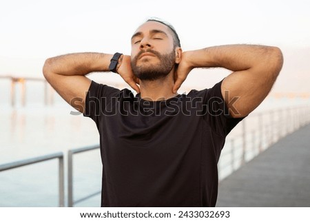 Sporty man in black t-shirt closes his eyes touching neck, having moment of peace, relieving from aching muscles discomfort and neckache after outdoor workout by the sea Royalty-Free Stock Photo #2433032693