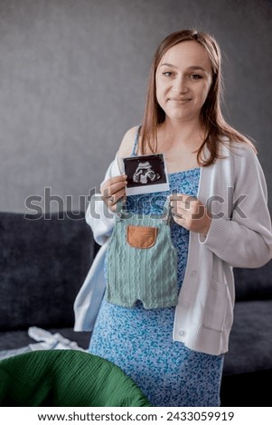 Beautiful pregnant woman looking a cute baby clothes and ultrasound picture at home. Anticipation of a child birth