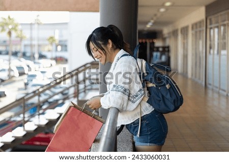 Horizontal photography of young adult woman traveling alone and going shopping, watching store sales while standing on the balcony, smiling and holding her bags