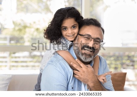 Happy loving Indian grandfather piggybacking sweet beloved preschool granddaughter at home, sitting on couch, enjoying kids hug, looking at camera, smiling, laughing Royalty-Free Stock Photo #2433065335