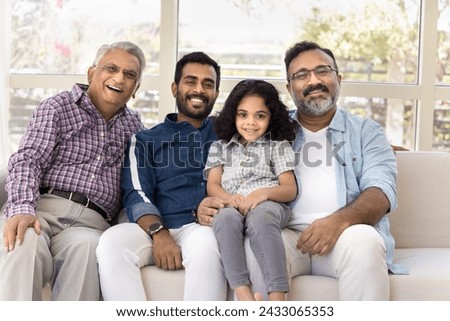 Cheerful kid and men of four family generations sitting close together on sofa, laughing, hugging little child with, looking at camera for portrait. Girl, father, grandpa, great grandfather portrait