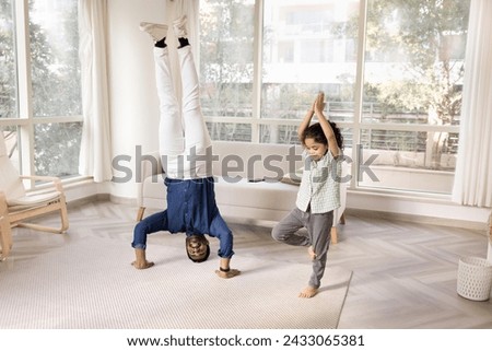 Happy active Indian dad and little daughter kid practicing yoga at home, exercising together, enjoying activity, active hobby, talking in headstand asana and tree pose Royalty-Free Stock Photo #2433065381