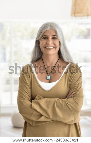 Happy confident senior woman with long naturally grey hair standing in home interior, looking at camera with crossed arms, smiling, posing for head shot vertical front portrait