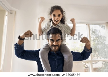 Cheerful strong Indian dad lifting little daughter kid on shoulders, making power hands gestures, showing fists, muscles, looking at camera with martial scream, playing active games Royalty-Free Stock Photo #2433065489