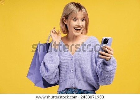 Beautiful, excited woman holding shopping bag and using mobile phone, paying, ordering, using mobile application. Attractive female student isolated on yellow background. Online shopping concept Royalty-Free Stock Photo #2433071933