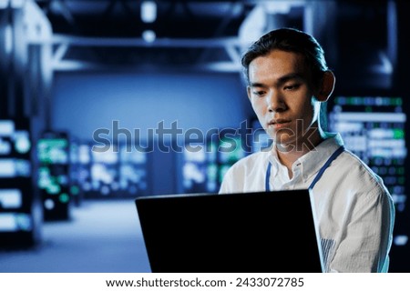 Skilled programmer in data center uses laptop to prevent system overload during peak traffic periods. Experienced professional in server room ensuring enough network bandwidth for smooth operations