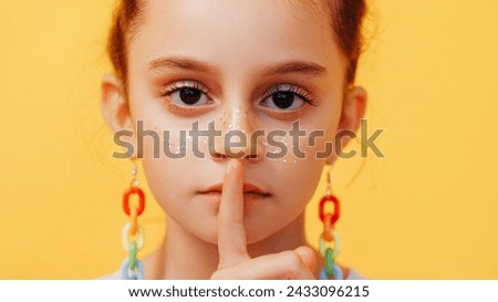 Kid secret. Hush child. Keep quiet. Closeup of worried serious little girl with fake glitter freckles in colorful earrings finger near mouth isolated on yellow background.