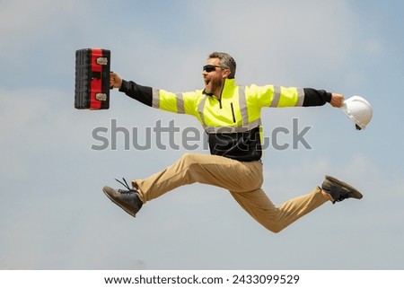 Crazy builder excited jump on site construction. Excited builder construction worker in a safety helmet jumping in front of the trucks. Excited crazy builder man in helmet jump outdoor. Royalty-Free Stock Photo #2433099529