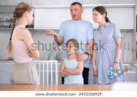 Displeased mother and father reprimanding eldest teenage daughter while standing with her younger sister in home kitchen. Conflict between parents and children Royalty-Free Stock Photo #2433106929