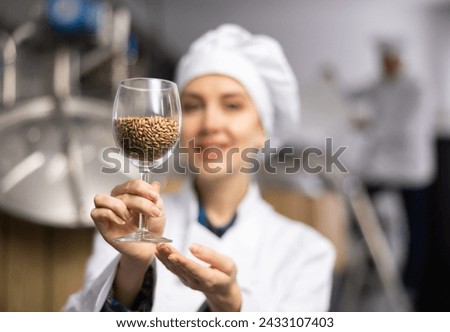 Skilled satisfied female brewer in white uniform demonstrating high quality malted barley grain in glass while standing at brewery, selective focus on glass with malt Royalty-Free Stock Photo #2433107403