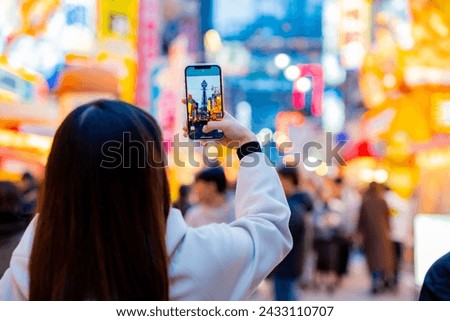Asian female tourist Traveling and having fun. And she was taking photo with a cell phone camera at Night street with many restaurant around Tsutenkaku Tower in Shinsekai district of Osaka, Japan.
