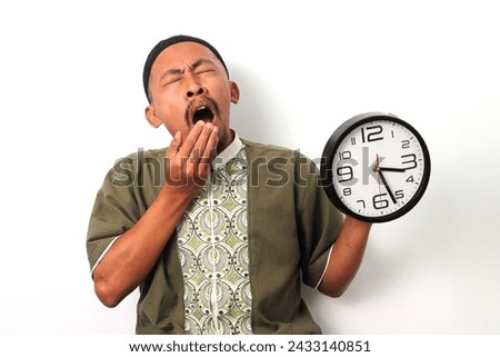 Sleepy Indonesian Muslim man in koko and peci stifles a yawn while checking a clock. He has just woken up to prepare for the suhoor meal before the Ramadan fast begins. Isolated on a white background