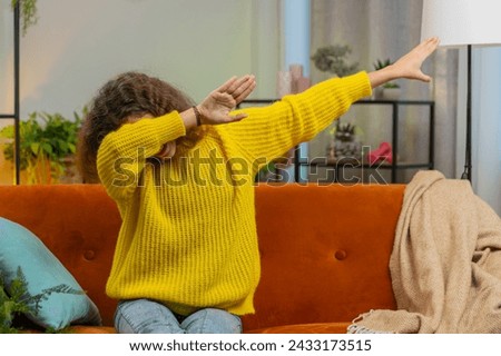 Trendy cheerful positive 14-15 years girl having fun dancing, moving to rhythm dabbing raising hands making dub dance gesture. Teenager Caucasian child kid at home in play living room sitting on couch Royalty-Free Stock Photo #2433173515
