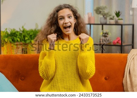 Happy child girl shouting, celebrating success, winning, goal achievement good news, lottery luck victory, watching TV championship at home apartment. Teen 14-15 years kid in living room sits on couch Royalty-Free Stock Photo #2433173525