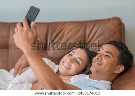 Happy young couple laying on the sofa at home and have fun using mobile phone to take selfie picture for social media pages account. Modern people man and woman living together in relationship love