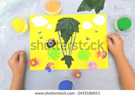 Child making card from paper, plasticine and natural leaves. Air balloon, clouds, birds, flowers... Inspiration for children. Plasticine and paper art children project. Travel holiday concept