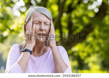 A close-up photo of an older gray-haired woman standing on the street in the park and holding her head with her hands, grimacing in pain, feeling strong pressure.