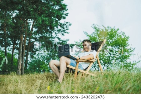 A man on a folding wooden chair sits in nature and works remotely online on a laptop. The concept of recreation in nature alone.