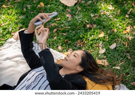 A carefree and happy Asian businesswoman is using her smartphone while lying on a picnic mat in a park, relaxing after work. people and lifestyle concepts