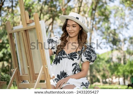 An attractive Asian female freelance artist is painting a picture on a canvas easel in a green park on a bright day. leisure and hobby concept