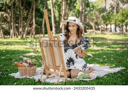 A charming Asian woman in a cute dress is painting a picture on a canvas easel while picnicking in a green park on the weekend. leisure and hobby concept