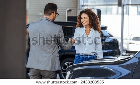Happy salesman congratulating his female customer for buying a new car in a showroom. Woman buying a new car in car dealership. Handshake and good deal 