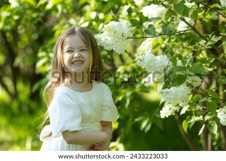 Cute little girl in white dress laughing in spring park near lilac. child smells lilac