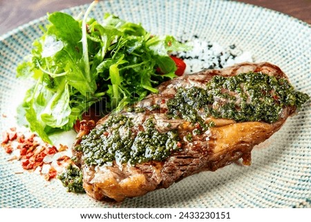 A perfectly grilled steak, adorned with a green herb sauce, rests beside a fresh green salad on a textured plate