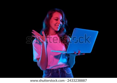 Smiling beautiful young woman holding laptop and talking via online video call with friends and colleagues against black studio background in neon light. Concept of youth, lifestyle, fashion, emotions