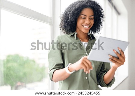 Optimistic happy African-American attractive young woman with a tablet in a bright room, engaged in online communication or leisure reading Royalty-Free Stock Photo #2433245455