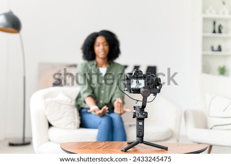 With a wave and a smile, an African-American woman engages her online audience, creating an inviting atmosphere in her vlog from a minimalist living space Royalty-Free Stock Photo #2433245475