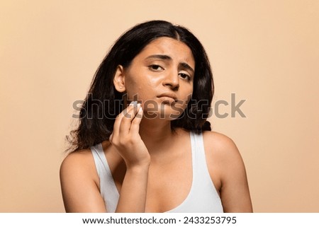 Upset Young Indian Woman Demonstrating Problems On Her Skin, Worried Eastern Female Touching Face And Looking At Camera, Stressed Lady Standing Isolated On Beige Studio Background, Copy Space Royalty-Free Stock Photo #2433253795