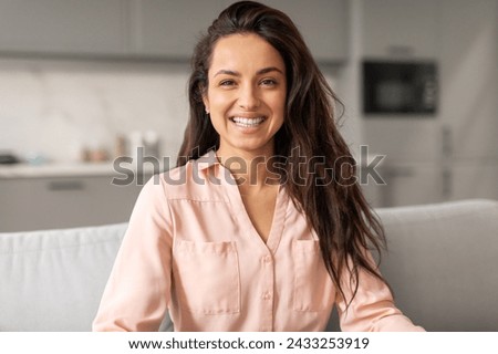 Vivacious woman with flowing hair and pastel shirt beams with warm, engaging smile while comfortably seated on chic grey sofa at home interior Royalty-Free Stock Photo #2433253919