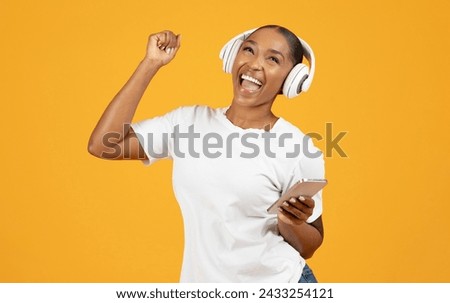 Millennial black woman in wireless headset with smartphone enjoys tunes on yellow studio background, dancing to music and having fun. Concept of modern technology and leisure